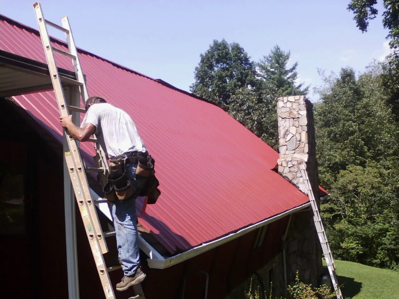 Roofer on a ladder beside a red metal roof and stone chimney — Soffit & Fascia Repair by Roofing Brothers in Hogansville